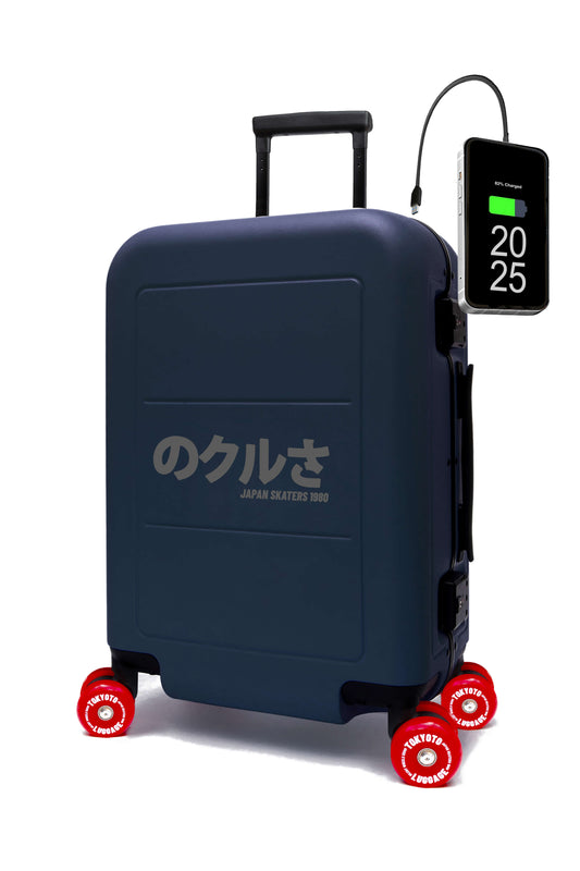 A dark navy blue hard-shell suitcase with red skate wheels and Japanese text on the front, shown on a white background with a smartphone charging cable plugged into its side.