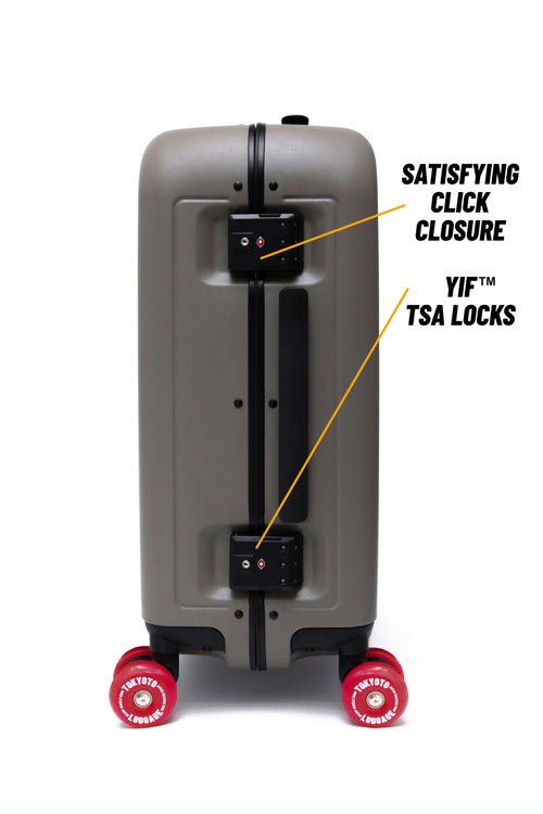 A gray hard-shell suitcase shown from the back, featuring black TSA lock and click closure hardware with yellow callout lines and text reading 'SATISFYING CLICK CLOSURE' and 'YIF TSA LOCKS.' The suitcase has red wheels and is photographed on a plain white background.