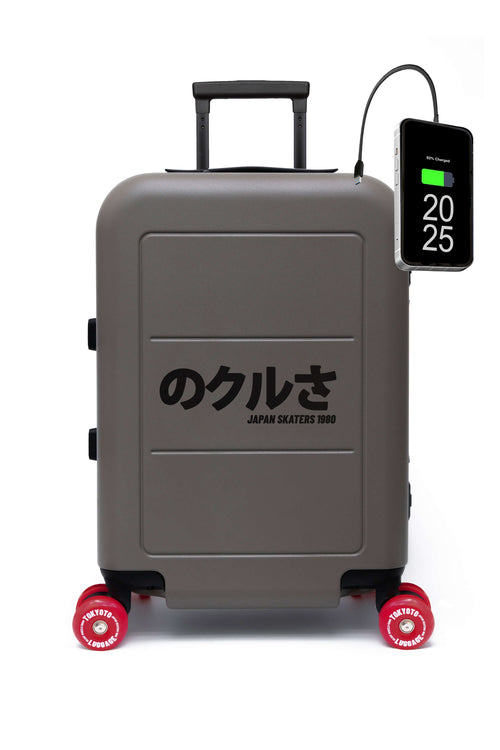 A gray hard-shell suitcase with red wheels and black Japanese text reading 'JAPAN SKATEERS 2025' on the front, shown on a white background with a phone charger plugged into it.