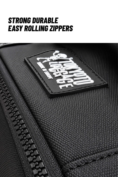 Close-up of a black textured fabric bag with a black zipper and a rectangular black patch featuring white embroidered text and a small cartoon figure. Text at the top reads 'STRONG DURABLE EASY ROLLING ZIPPERS'.