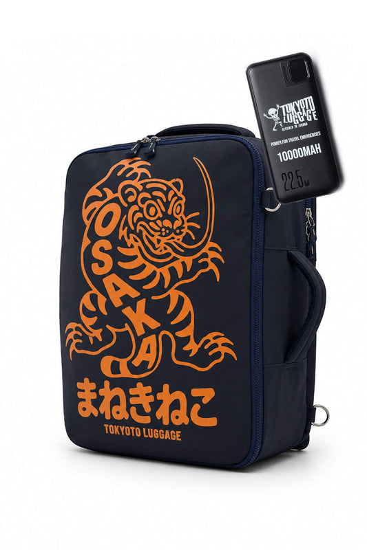 A navy blue soft-sided luggage bag with a large orange Tokyo Raccoon graphic and Japanese text, shown on a white background with a black phone case attached to the side.